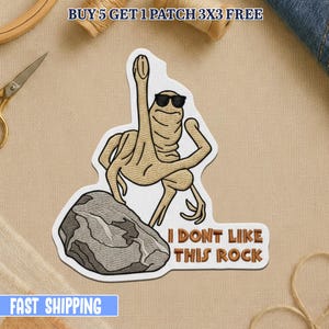 May include: Embroidered patch featuring a tan alien-like figure wearing sunglasses, flexing, and standing on a gray rock. The text "I DON'T LIKE THIS ROCK" is below the figure. The patch is on a white background.