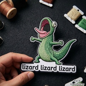 May include: Embroidered patch featuring a green lizard with an open mouth and pink tongue. The patch has the words "lizard lizard lizard" in a white rectangle. The patch is held by a hand, with spools of thread and a thimble in the background.