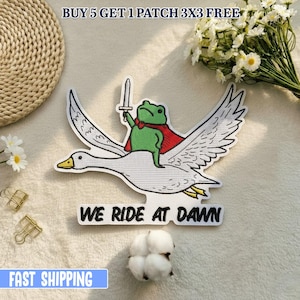Op de afbeelding: Geborduurde patch met een groene kikker in een rode cape, die een zwaard vasthoudt en op een witte gans rijdt. De patch heeft de tekst "WE RIDE AT DAWN". De patch is wit, groen, rood en geel.