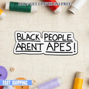 Peut inclure: Un écusson noir et blanc avec le texte "BLACK PEOPLE AREN'T APES!" dans un style manuscrit audacieux. L'écusson est entouré de bobines de fil et de boutons.