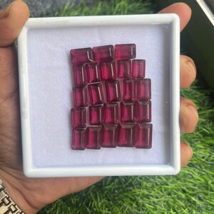 May include: A collection of rectangular, deep red gemstones displayed in a white tray. The gems are arranged closely together, showcasing their polished surfaces and rich color. The tray is held against a background of green grass.
