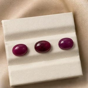 May include: Three oval-shaped, polished ruby gemstones displayed on a white, textured surface. The rubies vary in shade from deep red to a lighter, purplish-red. The gemstones are set against a neutral background.