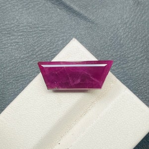 Puede incluir: Una piedra preciosa facetada de color rosa intenso con forma de trapecio. La piedra preciosa está colocada sobre una superficie blanca.
