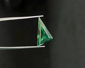 May include: A vibrant green gemstone with a triangular cut, held by silver-toned metal tweezers. The stone features multiple facets that reflect light, creating a sharp, geometric appearance with deep emerald hues and bright lime highlights.