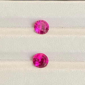 May include: Two round, faceted, hot pink gemstones are displayed against a white background. The gems have a brilliant cut, reflecting light. The gemstones are likely rubies or pink sapphires, perfect for jewelry making.