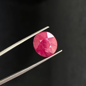May include: A deep red ruby gemstone with a faceted cut, held by tweezers against a black background.