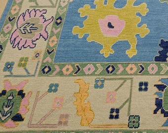 May include: A blue, beige, pink, green, and yellow patterned rug with a fringed edge. The rug has a floral design with a central motif of a stylized flower.