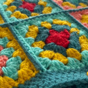 Pu&ograve; includere: Primo piano di una coperta all'uncinetto con una variet&agrave; di colori, tra cui turchese, giallo, rosso e rosa. La coperta &egrave; composta da quadrati, e ogni quadrato ha un motivo diverso. C'&egrave; un bottone giallo sulla coperta.