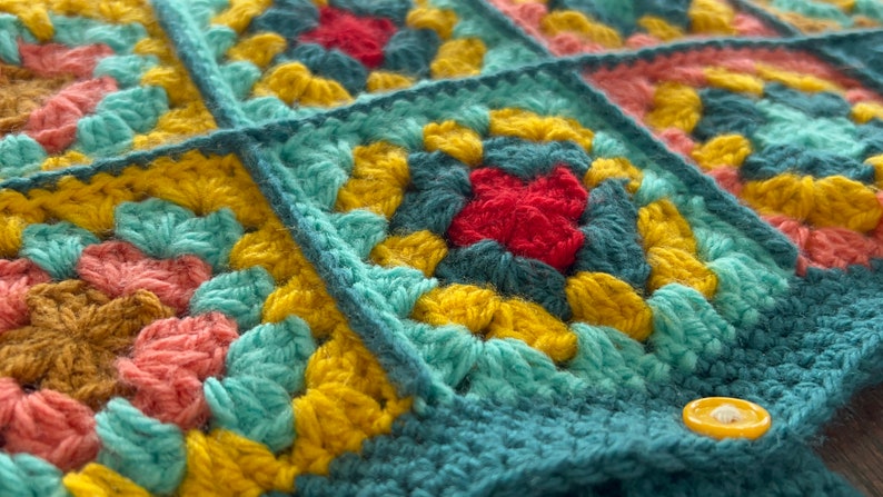 Pu&ograve; includere: Primo piano di una coperta all'uncinetto con una variet&agrave; di colori, tra cui turchese, giallo, rosso e rosa. La coperta &egrave; composta da quadrati, e ogni quadrato ha un motivo diverso. C'&egrave; un bottone giallo sulla coperta.