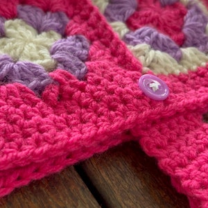 Pu&ograve; includere: Una coperta rosa all'uncinetto con un motivo viola e bianco. La coperta ha un bottone viola.