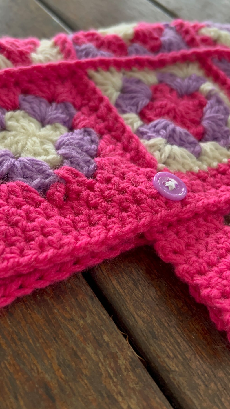 Pu&ograve; includere: Una coperta rosa all'uncinetto con un motivo viola e bianco. La coperta ha un bottone viola.