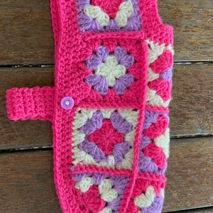 Pu&ograve; includere: Un maglione per cani in crochet rosa con un motivo viola e bianco. Il maglione ha una chiusura a bottone e una cinghia.