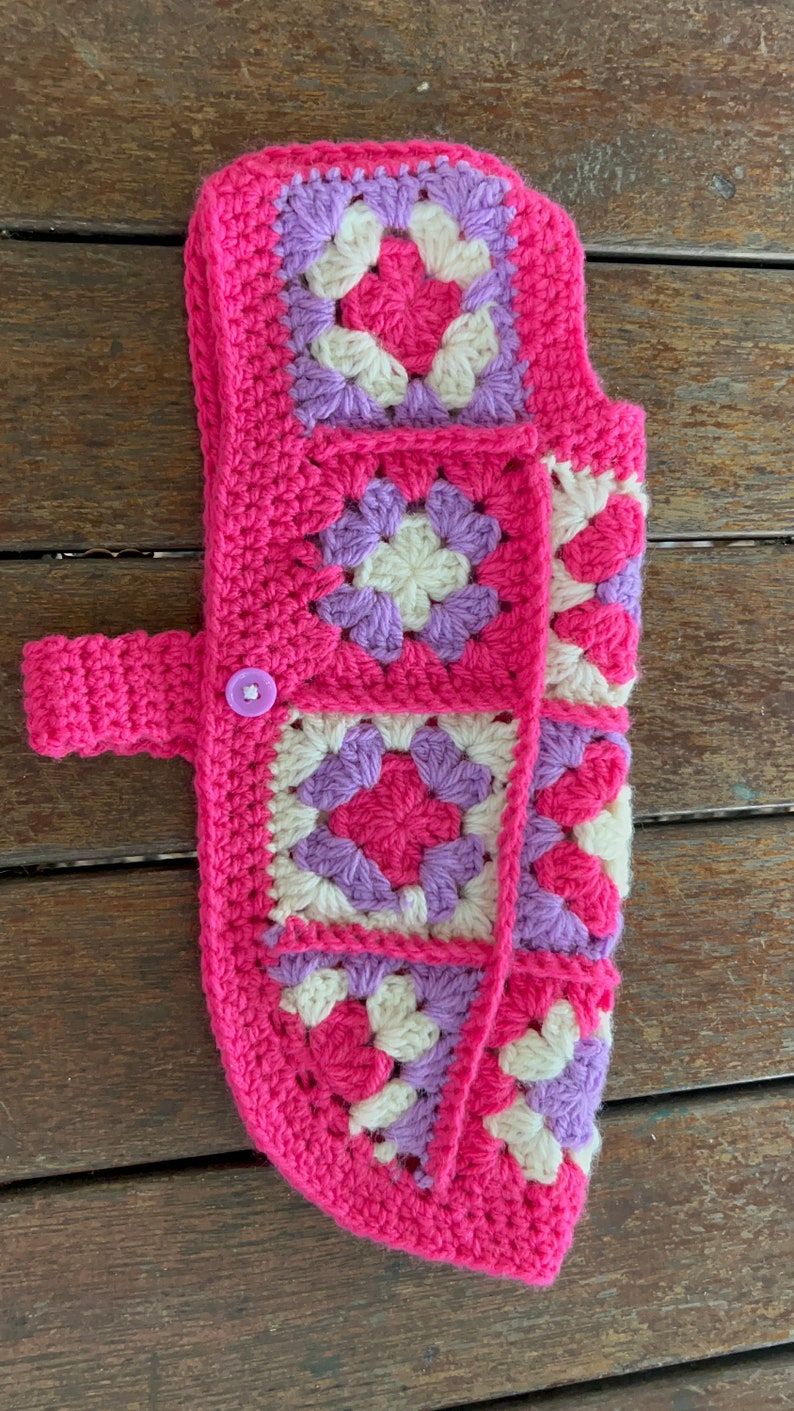 Pu&ograve; includere: Un maglione per cani in crochet rosa con un motivo viola e bianco. Il maglione ha una chiusura a bottone e una cinghia.