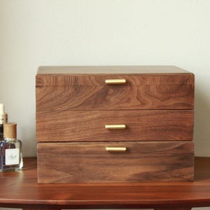 May include: A three-drawer wooden jewellery box with brass handles. The box is made of dark brown wood and sits on a matching wooden table. Several cosmetic products are arranged on the table next to the box.