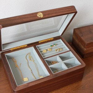 May include: A wooden jewelry box with a clear glass top, revealing compartments lined with light gray fabric. The box is open, displaying necklaces, earrings, and rings. The wood has a rich, dark brown finish, and the hardware is gold-toned.