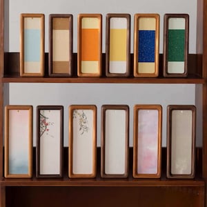Puede incluir: Una colección de marcos rectangulares de madera con varios insertos coloridos y texturizados, dispuestos en dos estantes. Los insertos presentan tonos de azul, naranja, amarillo y verde, así como diseños estampados y artísticos. Los marcos se exhiben sobre un fondo de madera oscura.