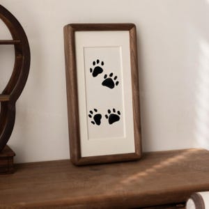 May include: A rectangular wall art piece featuring a dark brown wooden frame and a cream-colored mat board. The center of the artwork displays four black animal paw prints arranged in a vertical pattern on a white background.