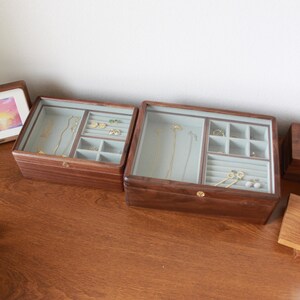 May include: Two wooden jewelry boxes with glass lids. The boxes are filled with necklaces, earrings, and rings. The interior is a light blue color. The boxes are sitting on a wooden surface.