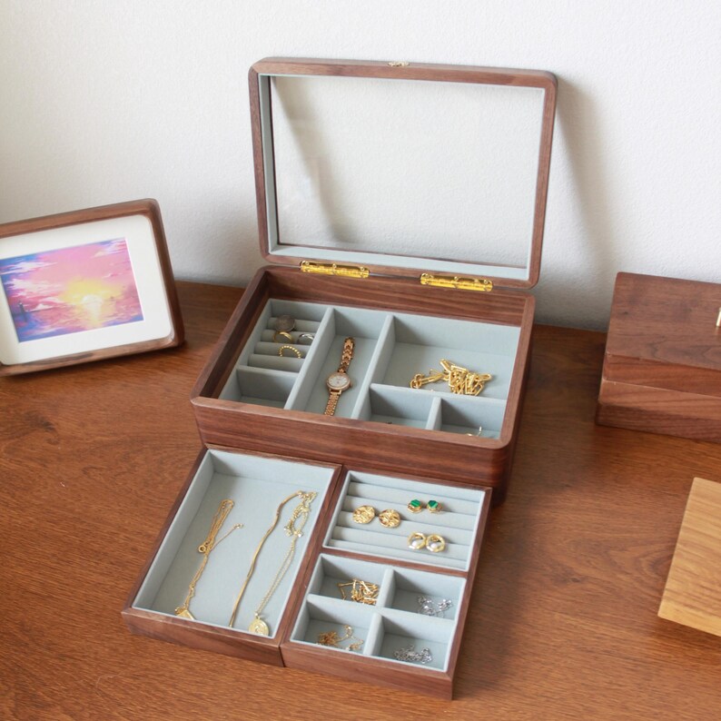 May include: A wooden jewelry box with a clear glass top, open to reveal multiple compartments lined with gray fabric. The box contains various pieces of jewelry, including necklaces, rings, earrings, and a wristwatch. The box is on a wooden surface.