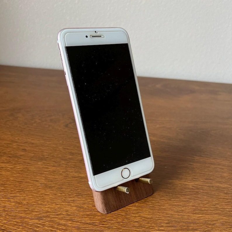 May include: A white smartphone with a black screen is propped up on a wooden stand with two brass accents. The stand is on a wooden surface.