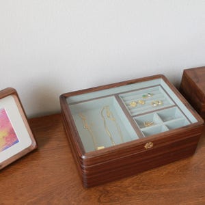 May include: A wooden jewelry box with a glass top, showcasing various compartments for rings, necklaces, and other accessories. The box is a rich brown color, with a light blue interior. Gold necklaces and rings are visible inside.