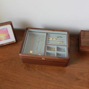 May include: A wooden jewelry box with a glass top, showcasing various compartments filled with gold necklaces and earrings. The box is a rich brown color and sits on a wooden surface, alongside a framed picture.