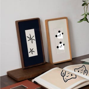May include: Two rectangular wooden frames stand upright. The left frame has a dark wood border, a navy blue mat, and a white insert with two black star-like drawings. The right frame has a light wood border, a white mat, and two panda illustrations.