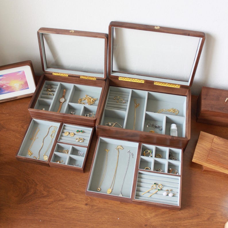 May include: Two wooden jewelry boxes with clear glass lids are open, revealing compartments filled with jewelry. The boxes are brown with gold-colored hardware. Smaller trays are pulled out, displaying necklaces, rings, and earrings. A framed picture is on the left.