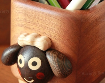 May include: A wooden pen holder with a sheep design. The pen holder is made of light and dark brown wood. The sheep has a dark brown face with light brown ears and a light brown cloud-shaped top. Pens and pencils are visible inside the holder.