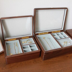 May include: Two wooden jewelry boxes with glass lids are open, revealing compartments lined with light blue fabric. Gold-colored clasps and hinges secure the lids. The boxes contain various gold necklaces, rings, and earrings.