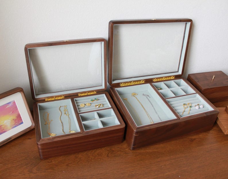 May include: Two wooden jewelry boxes with glass lids are open, revealing compartments lined with light blue fabric. Gold-colored clasps and hinges secure the lids. The boxes contain various gold necklaces, rings, and earrings.