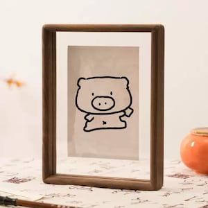 May include: A rectangular wooden picture frame with a clear glass front. It contains a black ink drawing of a cartoon pig on a light background. The frame has rounded corners.