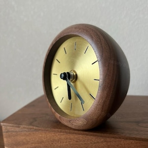 May include: A gold-toned clock face with black hands set in a dark brown wooden frame. The clock is a decorative piece for a home or office.