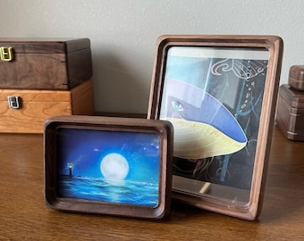 May include: Two wooden picture frames with different images. The frame on the left has a seascape with a lighthouse and a full moon. The frame on the right has a blue and yellow whale with a white background.