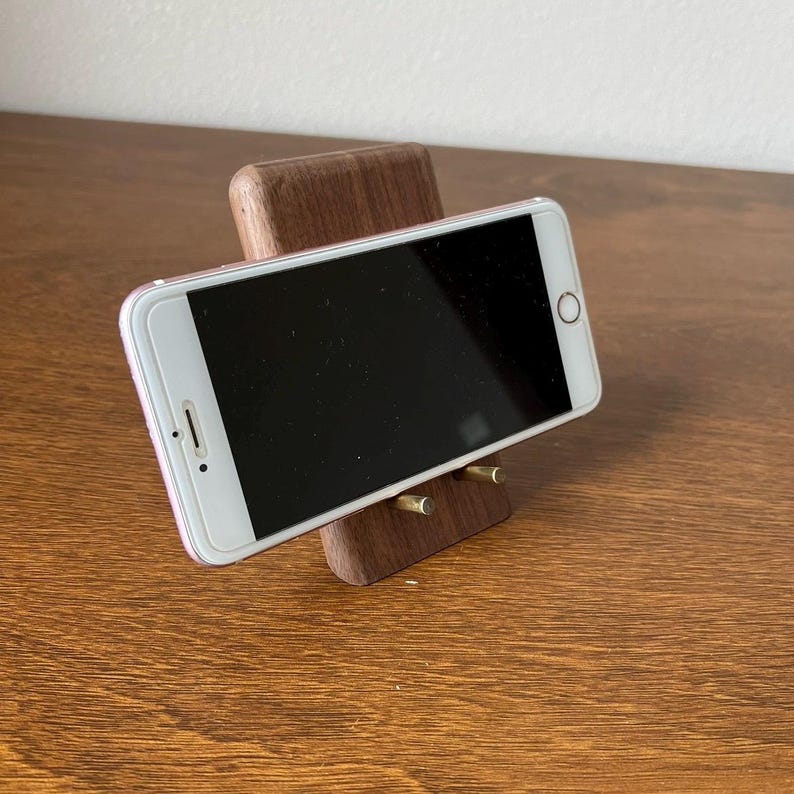 May include: A wooden phone stand with two brass pegs to hold a smartphone in place. The stand is made of dark wood and has a natural finish.