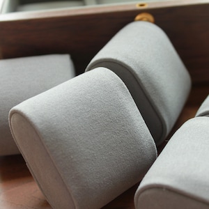 May include: Close-up of several grey watch pillows, likely for display or storage. The pillows are made of a soft, textured material and are arranged inside a wooden box. The pillows are cylindrical with rounded tops.