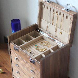 May include: A wooden jewellery box with a hinged lid and four drawers. The lid has a velvet lining with compartments for rings, earrings, and necklaces. The box has a brown wood finish and silver-toned hardware.