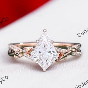 May include: Rose gold engagement ring with a large, kite-shaped diamond centre stone. The band features a delicate, intertwined design with smaller accent stones, creating a sparkling and elegant look. The ring is set against a white background.