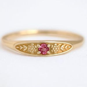 Puede incluir: Un anillo de oro con un diseño central ovalado. El anillo presenta una pequeña gema rosa redonda en el centro, flanqueada por motivos florales y de hojas. El anillo está sobre un fondo blanco.