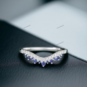 May include: A silver ring with a curved band and a row of small blue and white gemstones. The ring is set on a black surface.