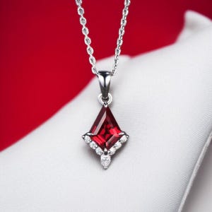 May include: A silver necklace featuring a red gemstone pendant. The pendant is a diamond shape with a central red stone and smaller white stones around the edges. The background is a solid red.