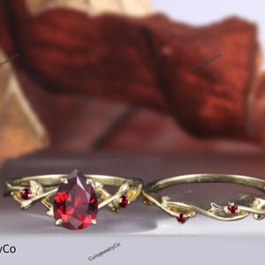 Può includere: Un paio di anelli d'oro con una pietra preziosa rossa a forma di pera incastonata al centro dell'anello più grande. L'anello più piccolo ha un design a vite con tre piccole pietre preziose rosse.