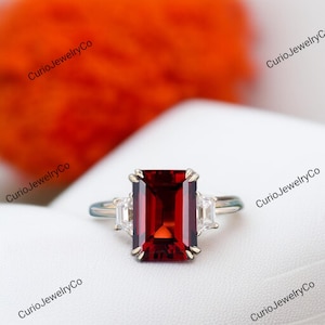 Puede incluir: Anillo plateado con una gran gema rectangular de color rojo intenso. Dos gemas claras rectangulares más pequeñas flanquean la piedra principal. El anillo se encuentra sobre un fondo blanco con un fondo naranja.