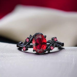 May include: Black ring with a large oval-cut red gemstone as the focal point. The ring features a floral design with smaller red accent stones and delicate leaf details. The ring is set against a soft, neutral background.