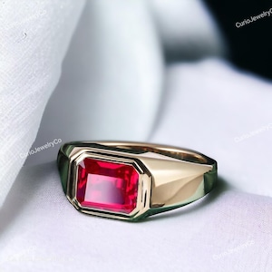 以下が含まれることがあります： 長方形の鮮やかな赤色の宝石が付いたゴールドカラーのリング。リングは、洗練された仕上げのクラシックな幾何学模様のデザインです。宝石はベゼル内にセットされており、大胆でエレガントな外観を作り出しています。リングは白い表面に表示されています。