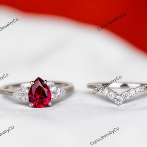May include: A two-piece ring set featuring a pear-shaped ruby gemstone ring and a curved band with small, round diamonds. The ruby is a deep red color, and the bands appear to be silver or white gold. The rings are set against a white background.