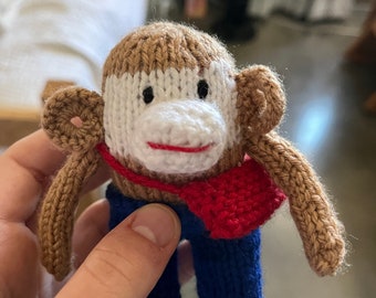 May include: A knitted monkey toy with brown fur, a white face, red scarf, and blue pants.
