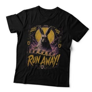 May include: A black short-sleeved t-shirt featuring a graphic print of a large rabbit in front of a yellow moon. Below the rabbit, a line of small figures runs across a hill. The text RUN AWAY! is printed in large yellow letters at the bottom.