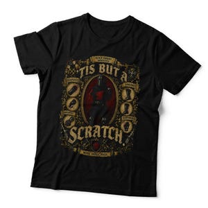 May include: A black short-sleeved t-shirt featuring a graphic print of a knight in armour. The design includes the text 'TIS BUT A SCRATCH' and 'NONE SHALL PASS' with illustrations of armour pieces and a central knight figure in a red oval frame.