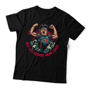 May include: A black short-sleeved t-shirt featuring a graphic of a muscular figure in a cap and suspenders with a red superhero emblem on the chest. The design includes skulls and the red text "NOT ALL HEROES WEAR CAPES" in a circular layout.